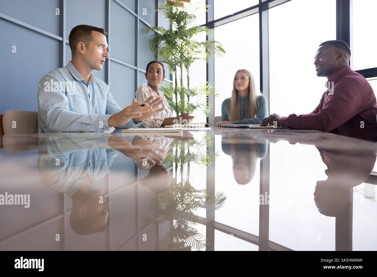 Un gruppo di quattro uomini d'affari che negoziano seduti attorno al tavolo da conferenza nella moderna sala meeting. Il team è impegnato in una discussione professionale, ascoltando i bot Foto Stock