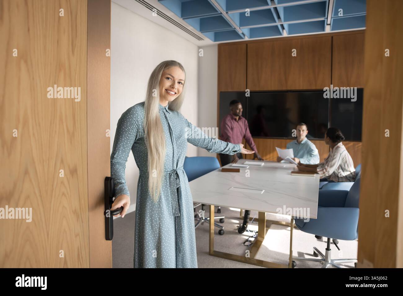 Una donna d'affari sorridente si trova davanti alla porta della moderna sala conferenze, facendo un gesto invitante e accogliendo partner o clienti per unirsi a una riunione, mentre il gruppo o Foto Stock