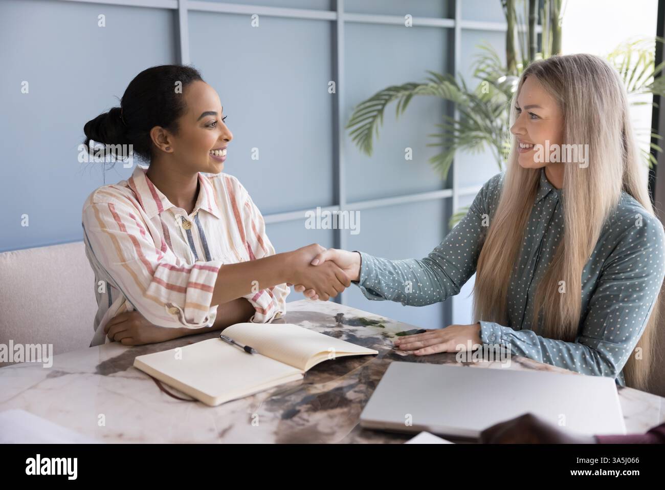 Due giovani donne d'affari multietniche che stringono la mano si siedono al tavolo di una conferenza, iniziano o finiscono una riunione formale, negoziano in ufficio, fanno affari, vendono Foto Stock