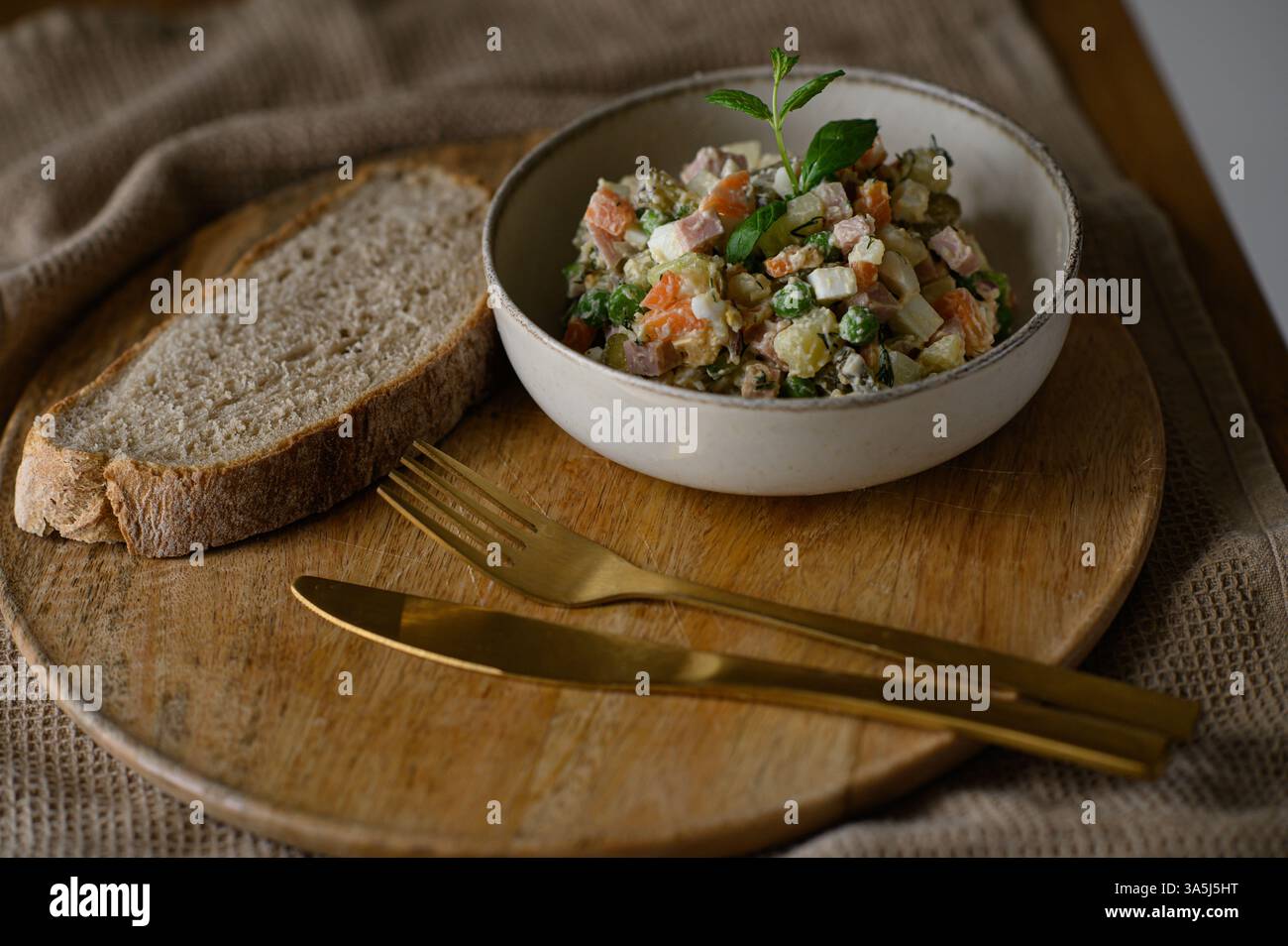 Un'insalata Olivier classica, cremosa e colorata, è servita elegantemente su un piatto di ceramica accanto a una fetta abbondante di pane integrale Foto Stock