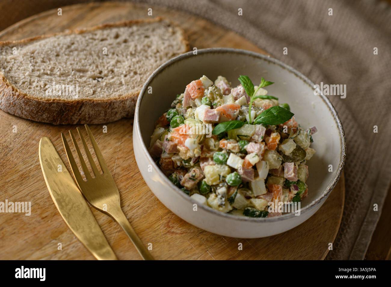 Un'insalata Olivier classica, cremosa e colorata, è servita elegantemente su un piatto di ceramica accanto a una fetta abbondante di pane integrale Foto Stock