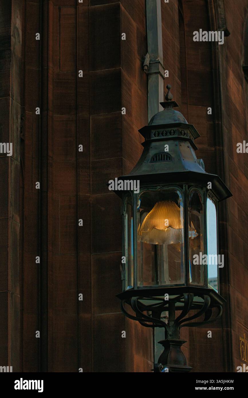 Una lanterna ornata è appesa al muro di un edificio, che emette un caldo bagliore. L'edificio ha una facciata in pietra con un design verticale a Manchester, Regno Unito. Foto Stock