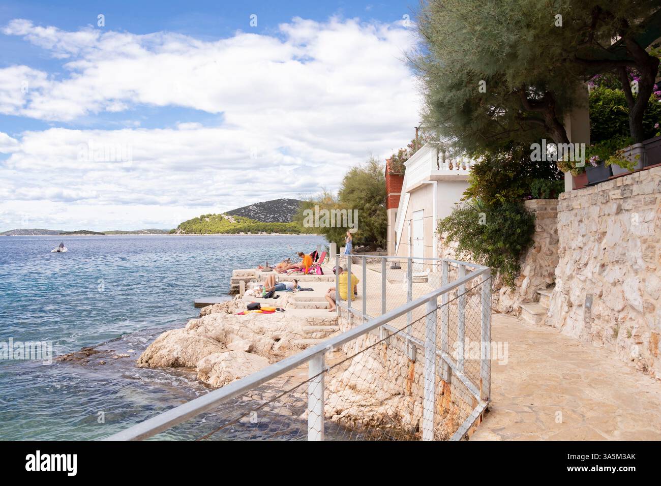 Tribunj, Croazia - 14 settembre 2024: Piccola e intima spiaggia rocciosa con poche persone che si rilassano di fronte alle case e al passaggio pedonale pavimentato Foto Stock
