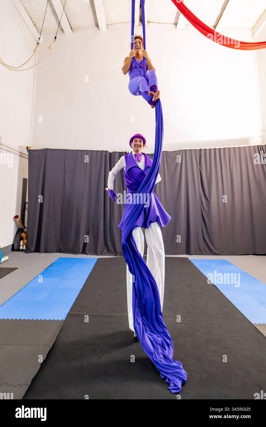 Due interpreti, una donna appesa alle sete aeree e un uomo su palafitte, praticano il loro circo recitano in palestra, mostrando forza, equilibrio e arte Foto Stock Due interpreti, una donna appesa alle sete aeree e un uomo su palafitte, praticano il loro circo recitano in palestra, mostrando forza, equilibrio e arte Foto Stock