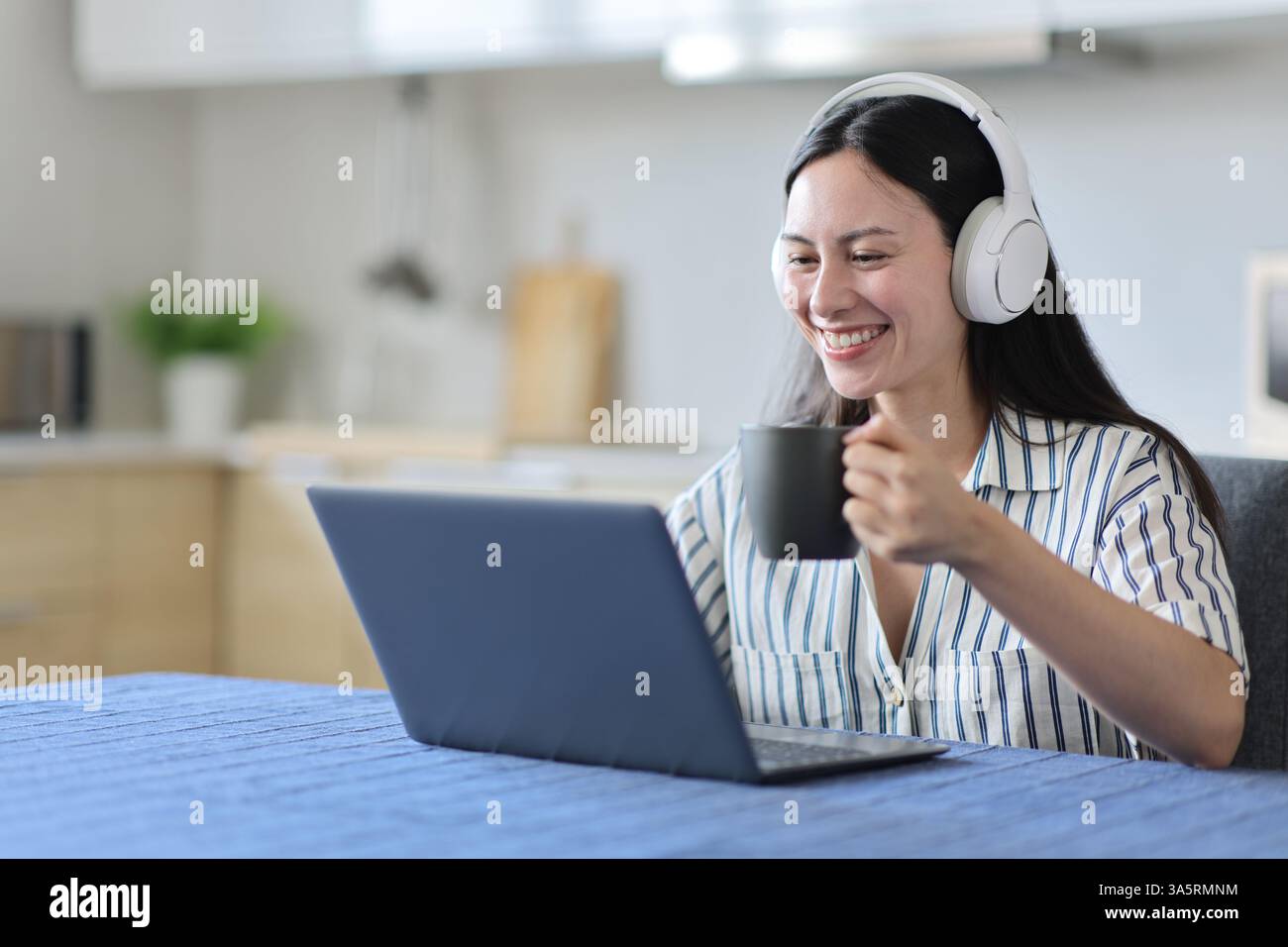 Felice donna asiatica che guarda i media sul portatile che beve in cucina a casa Foto Stock