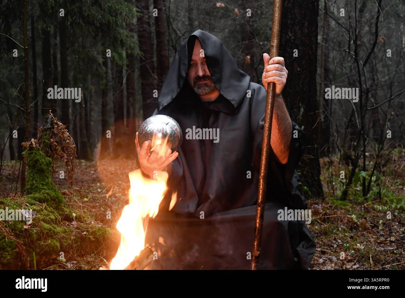 Un monaco dalle vesti nere siede accanto a un fuoco nella foresta e guarda in una palla di divinazione. Misticismo e magia, stregoni e streghe. Foto Stock
