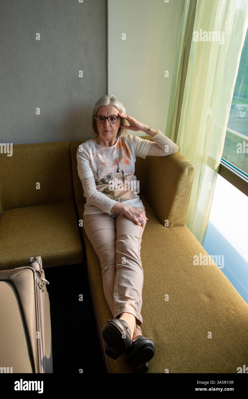 Una donna anziana siede comodamente su un divano vicino a una finestra luminosa, trasmettendo calma e introspezione in un tranquillo ambiente interno. Foto Stock