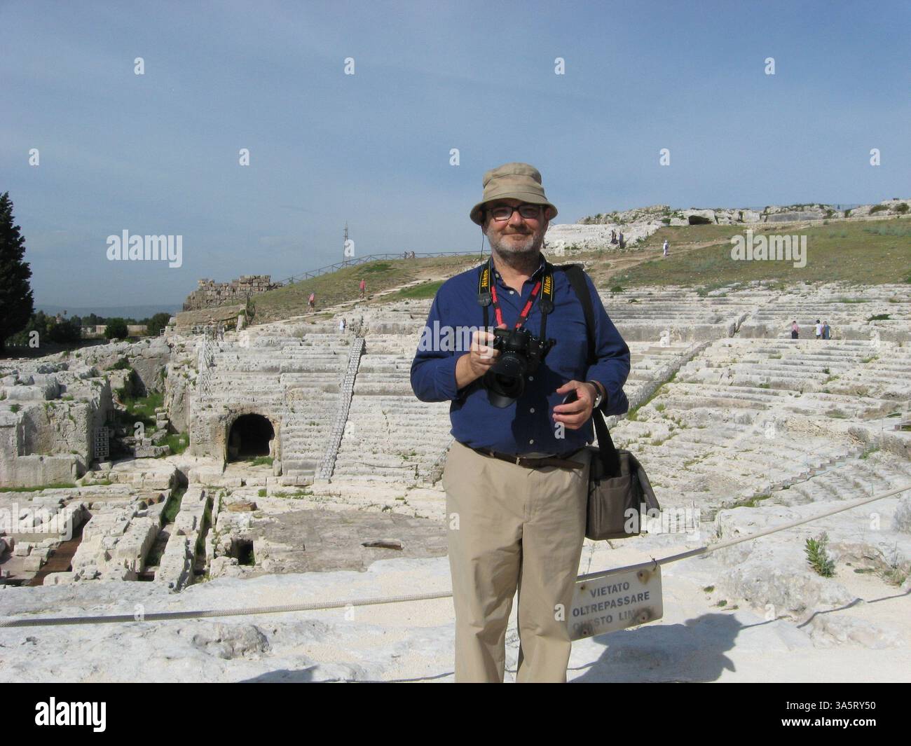 Un viaggiatore si trova in un anfiteatro storico, indossando un abbigliamento casual e tenendo in mano una macchina fotografica, sotto un cielo limpido e soleggiato, mostrando entusiasmo per la storia A. Foto Stock