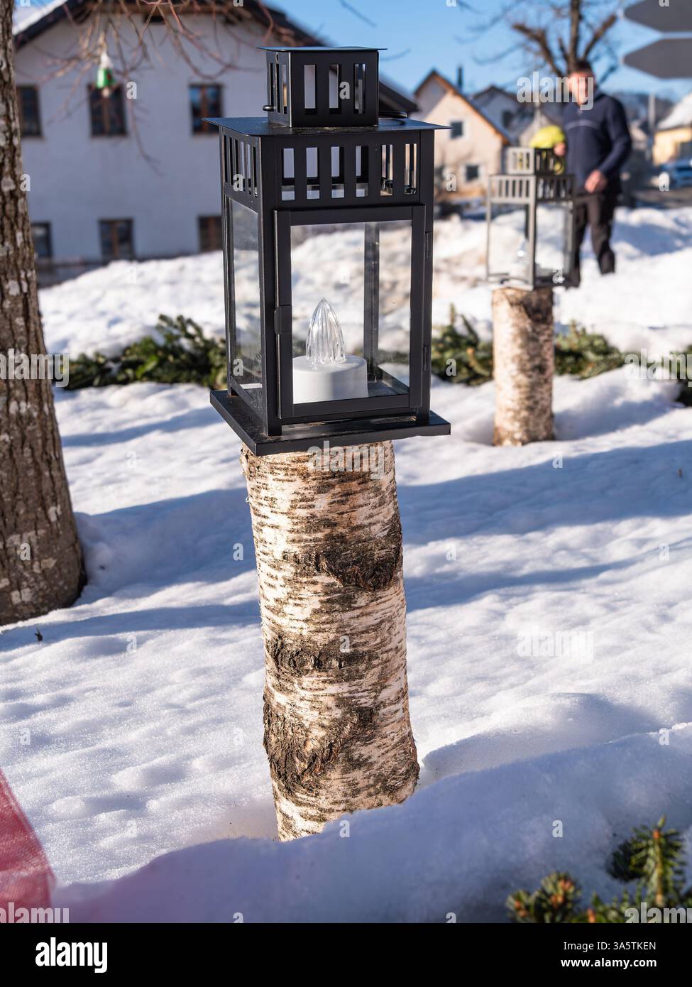 Una lanterna nera posizionata su un tronco di betulla in un ambiente all'aperto innevato, con una persona sfocata sullo sfondo. La scena include terreno innevato e. Foto Stock Una lanterna nera posizionata su un tronco di betulla in un ambiente all'aperto innevato, con una persona sfocata sullo sfondo. La scena include terreno innevato e. Foto Stock