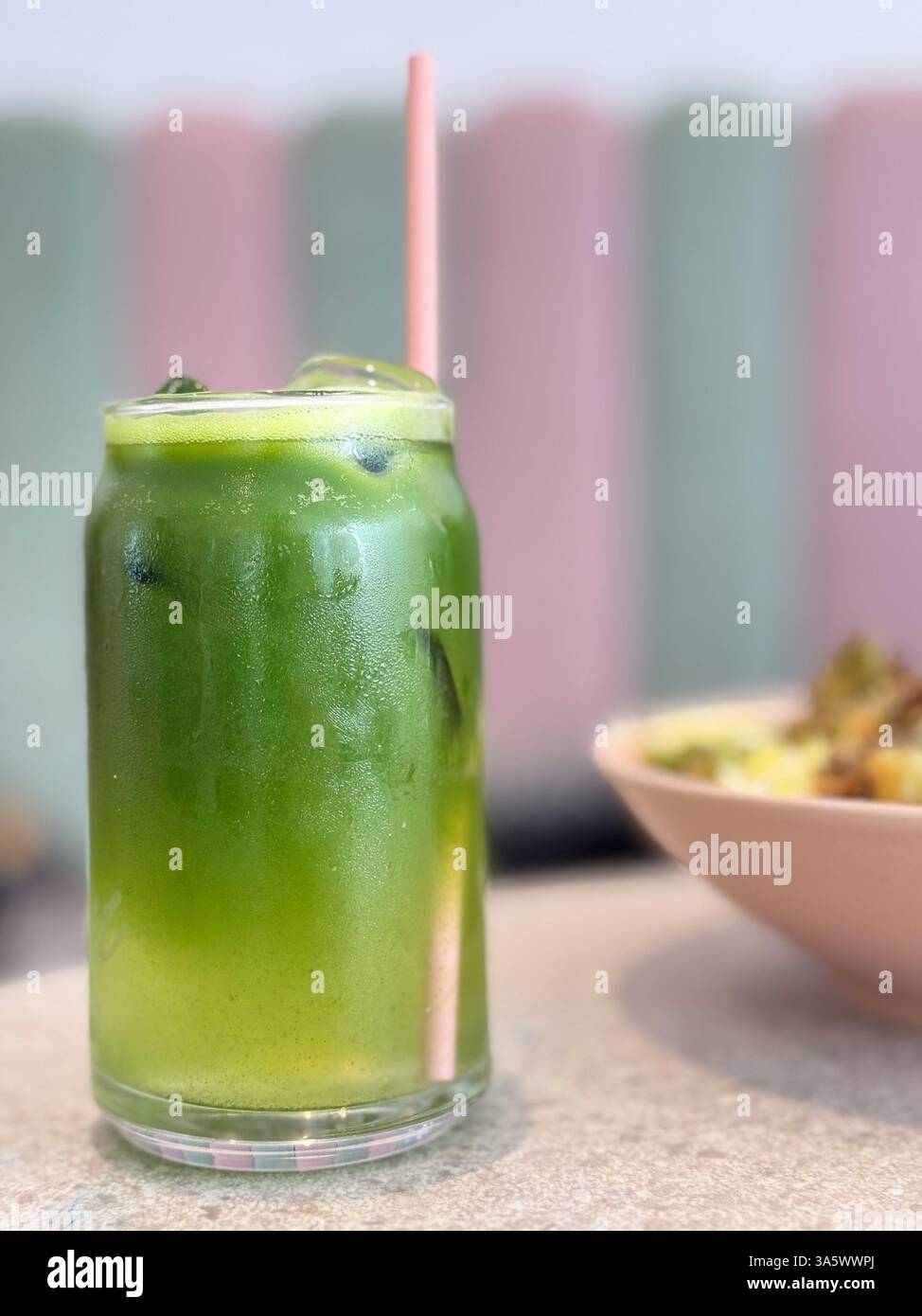 Un alto bicchiere di succo verde ghiacciato con una paglia rosa, seduto su un tavolo in un caffè dai toni pastello Foto Stock
