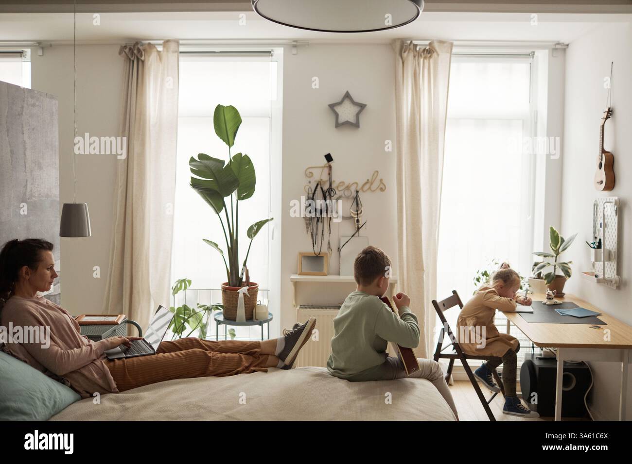 Visualizzazione laterale del ritratto di una madre single che lavora da casa e utilizza il computer portatile mentre è sdraiata sul letto con due bambini in un semplice interno Foto Stock