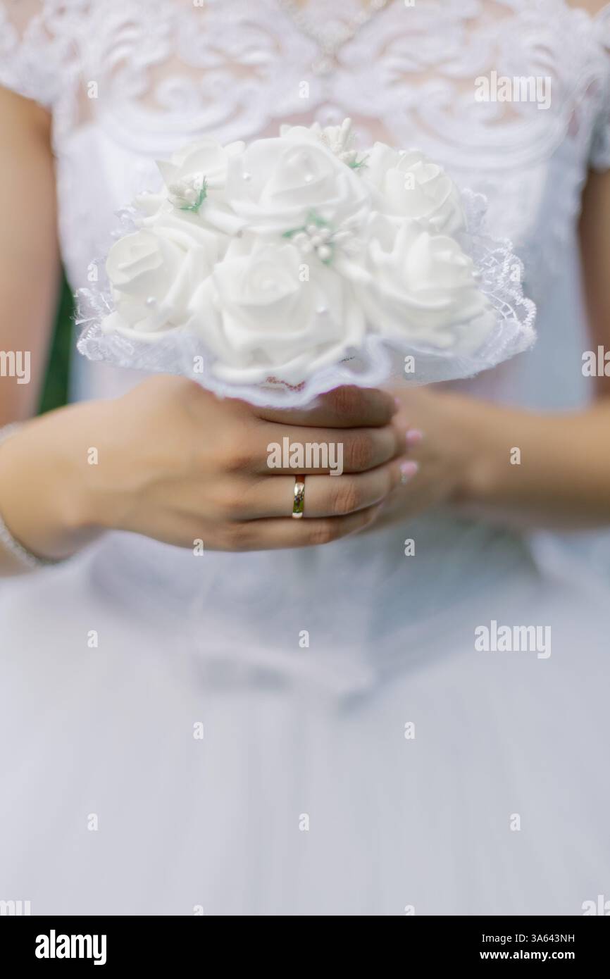 Una giovane e bella sposa in un abito da sposa bianco regala tra le mani un bouquet nuziale bianco su cui è realizzata una fede nuziale in oro Foto Stock