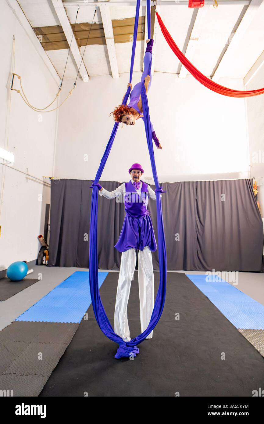 Due artisti del circo indossano costumi viola, eseguono acrobazie con sete aeree, una sospesa in aria e l'altra in piedi su palafitte Foto Stock Due artisti del circo indossano costumi viola, eseguono acrobazie con sete aeree, una sospesa in aria e l'altra in piedi su palafitte Foto Stock