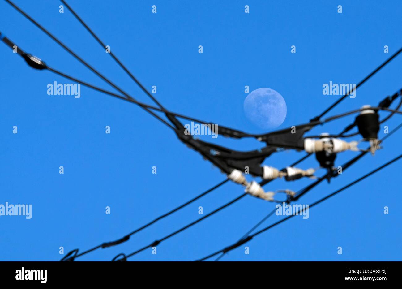 La luna sembra voltare sopra i cavi di alimentazione contro il cielo blu Foto Stock