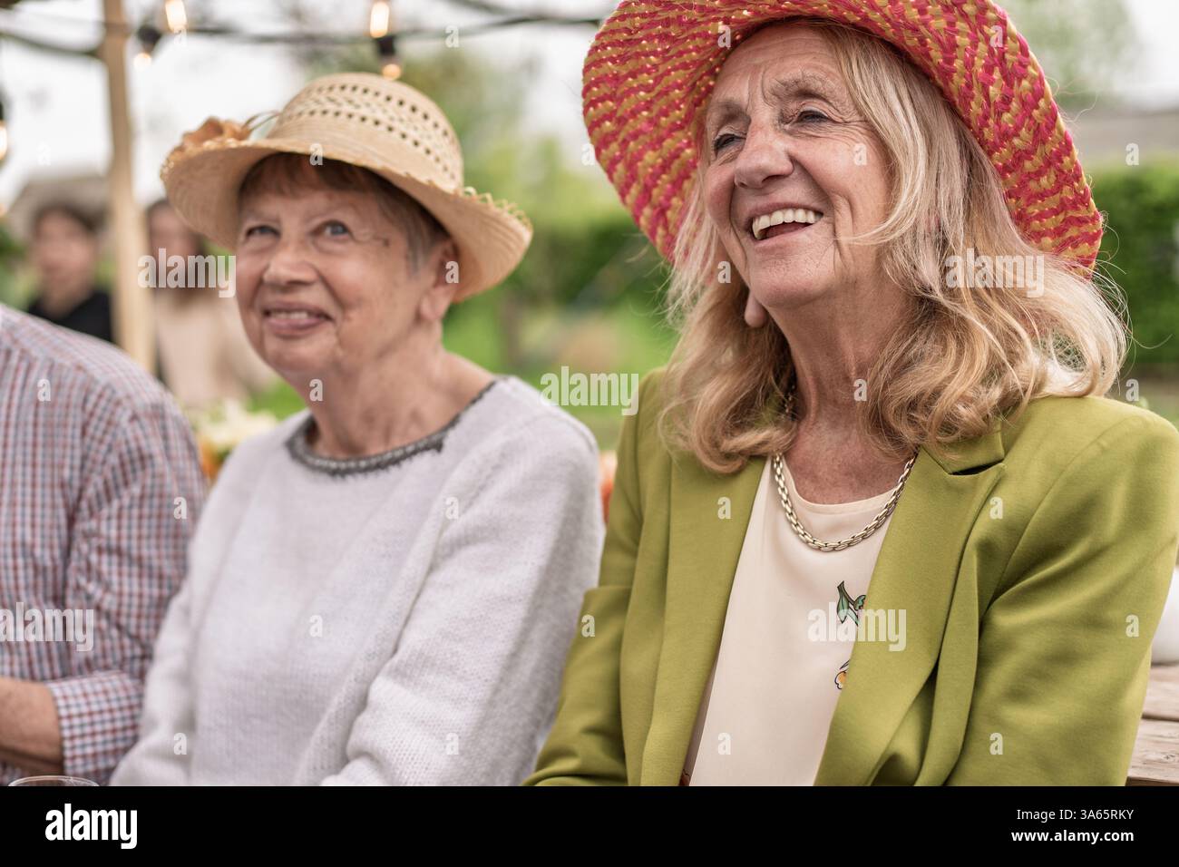 Due allegre donne anziane che si godono una festa all'aperto indossando cappelli di paglia e sorridendo, catturate in un'atmosfera luminosa e rilassata. Foto Stock