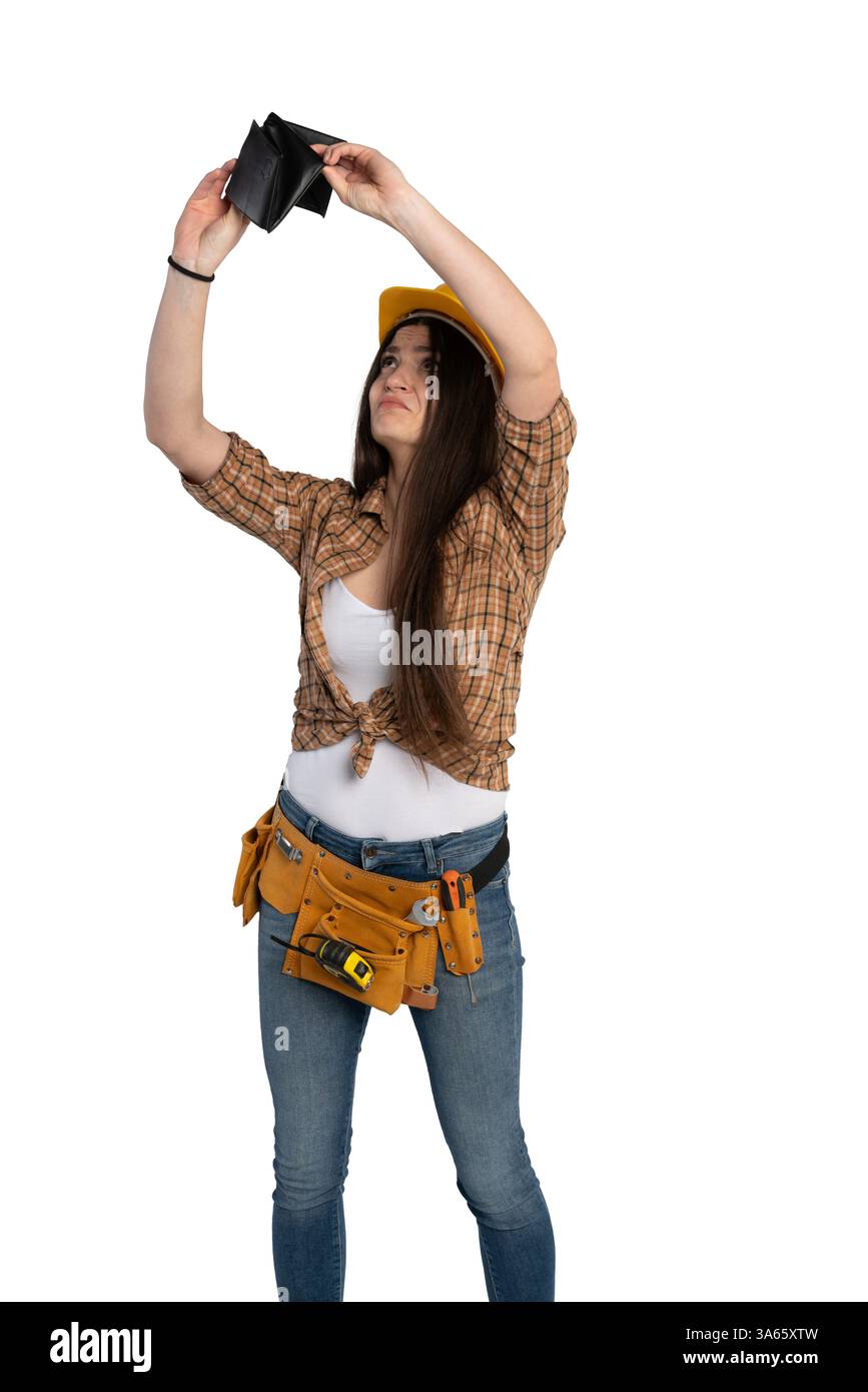 Una lavoratrice edile in una cintura portautensili e un elmetto che guarda un portafoglio vuoto, esprimendo preoccupazione finanziaria e incertezza. Foto Stock