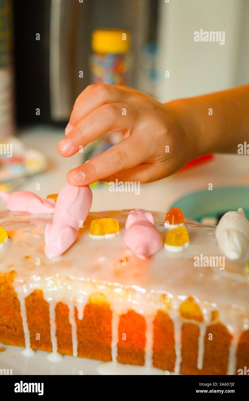 Questo scatto ravvicinato cattura la mano di un bambino posizionando delicatamente una decorazione bianca simile a caramelle su una torta di pane smerigliata. Foto Stock