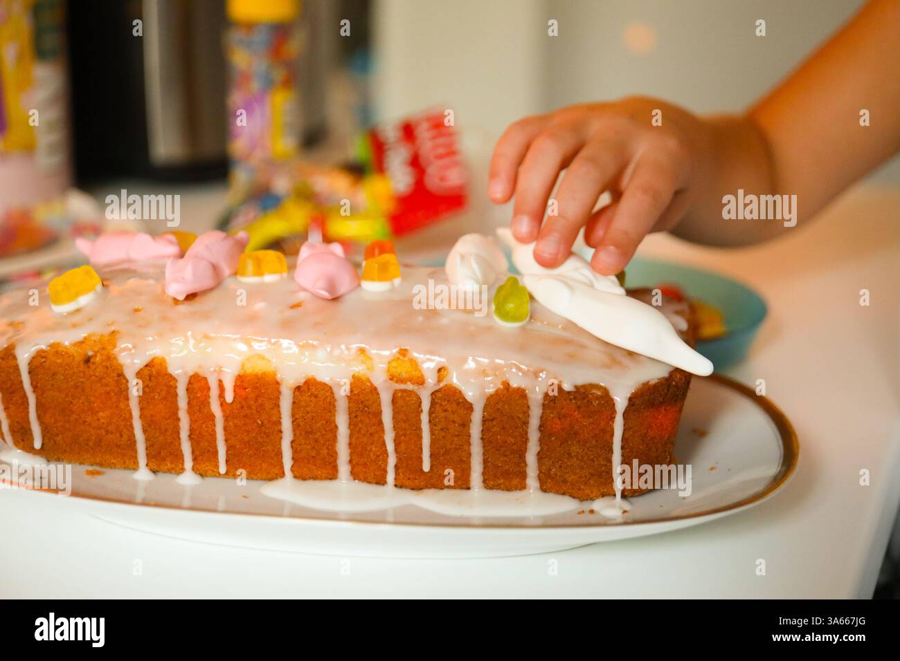 Questo scatto ravvicinato cattura la mano di un bambino posizionando delicatamente una decorazione bianca simile a caramelle su una torta di pane smerigliata. Foto Stock