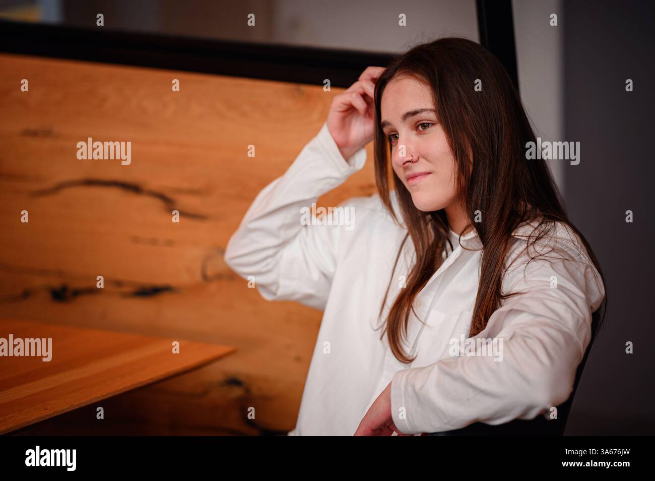 Una giovane donna con una camicia bianca si piega indietro con un sorriso delicato, toccando i suoi capelli. I caldi interni in legno del caffè aggiungono un tocco di intimità e relax Foto Stock