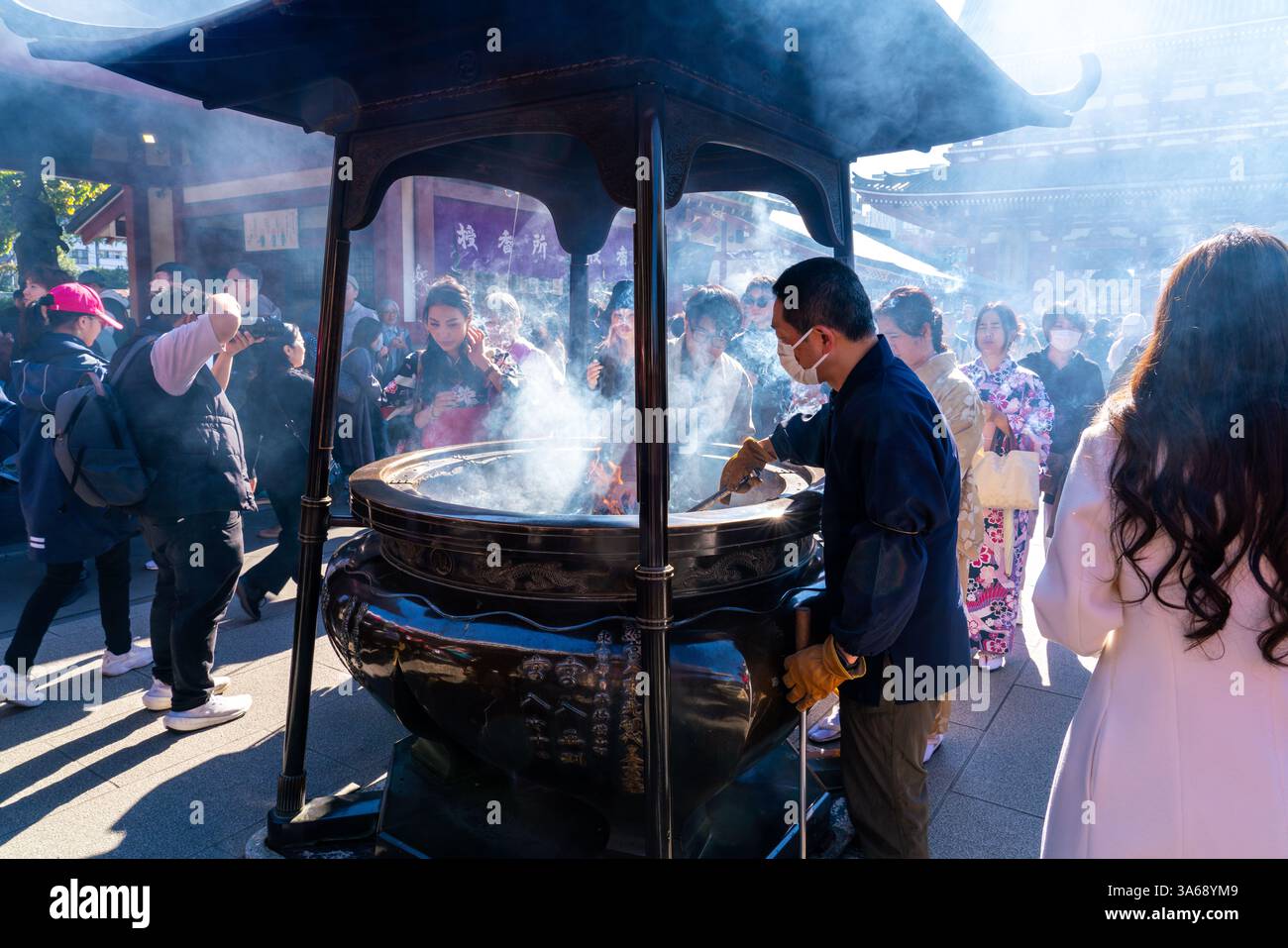 Uomo che accende incenso presso un bruciatore pubblico nel tempio Sensō-ji, Asakusa, Tokyo, circondato da visitatori. Foto Stock