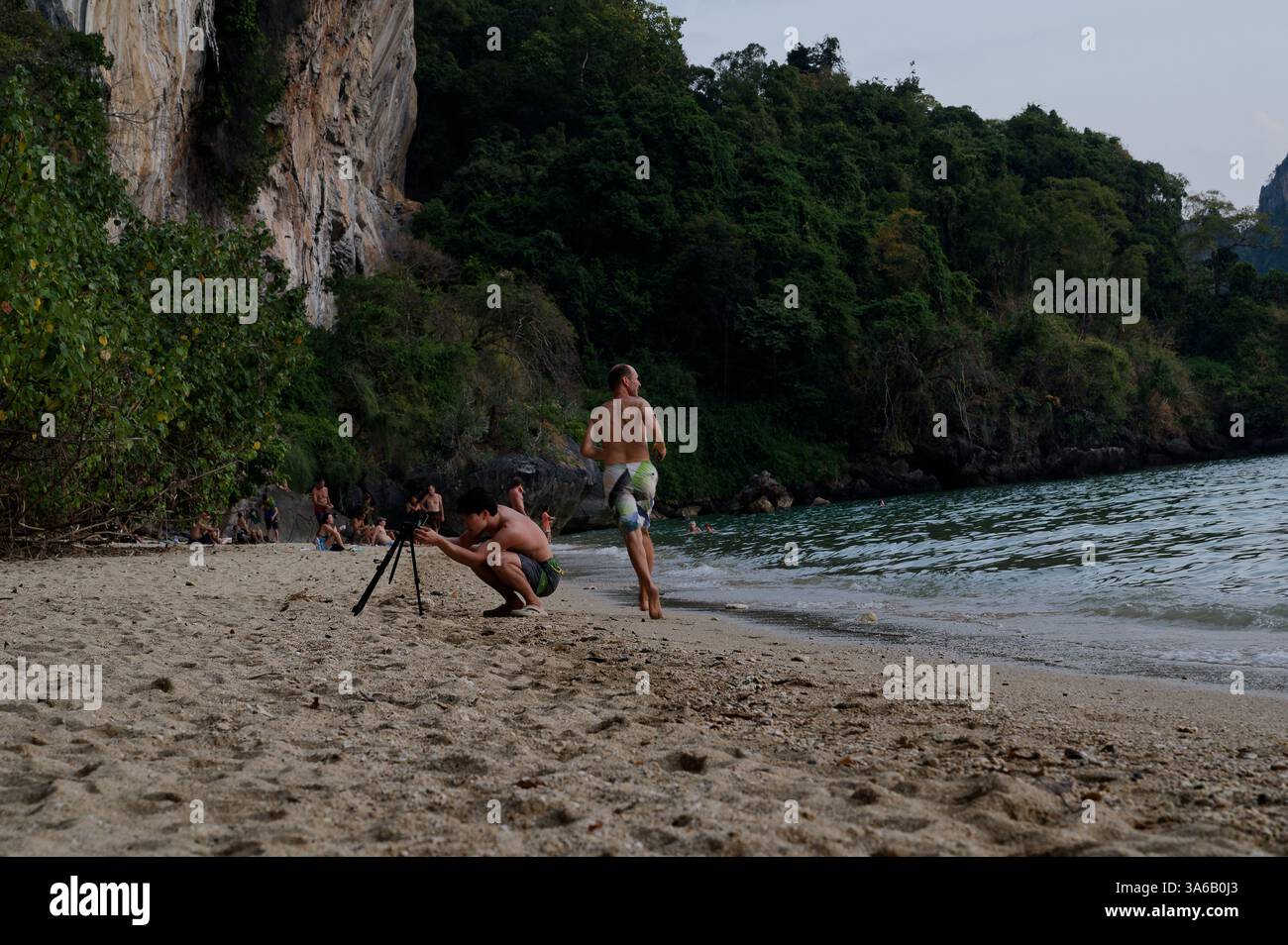 L'uomo prepara un treppiede sulla spiaggia di Tonsai come un altro sfreccia, con scogliere calcaree e vegetazione lussureggiante che si innalza sullo sfondo Foto Stock