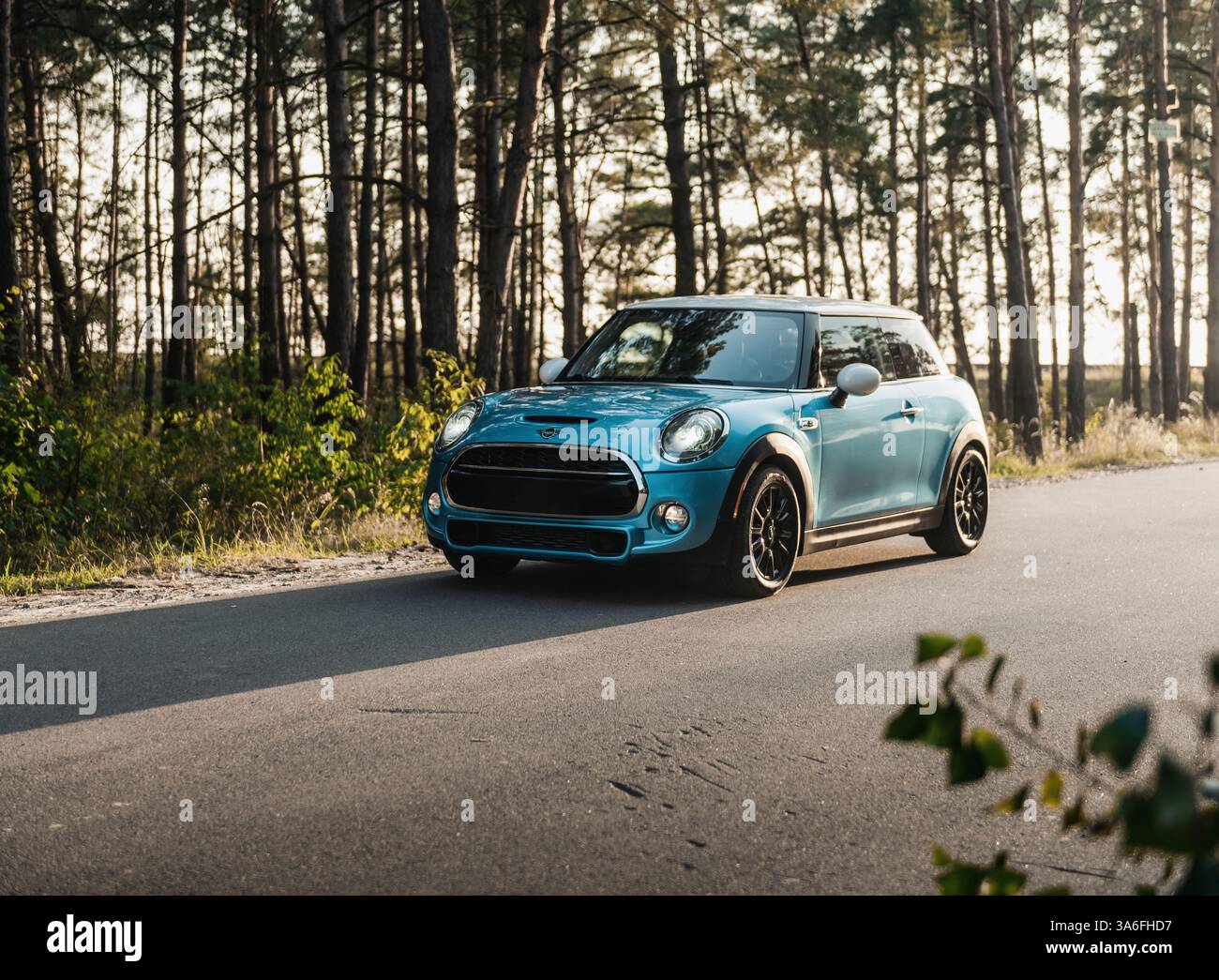 Blue Mini Cooper parcheggiata su una strada forestale in discesa. Vista frontale di tre quarti della berlina compatta britannica nel tardo pomeriggio. Foto Stock