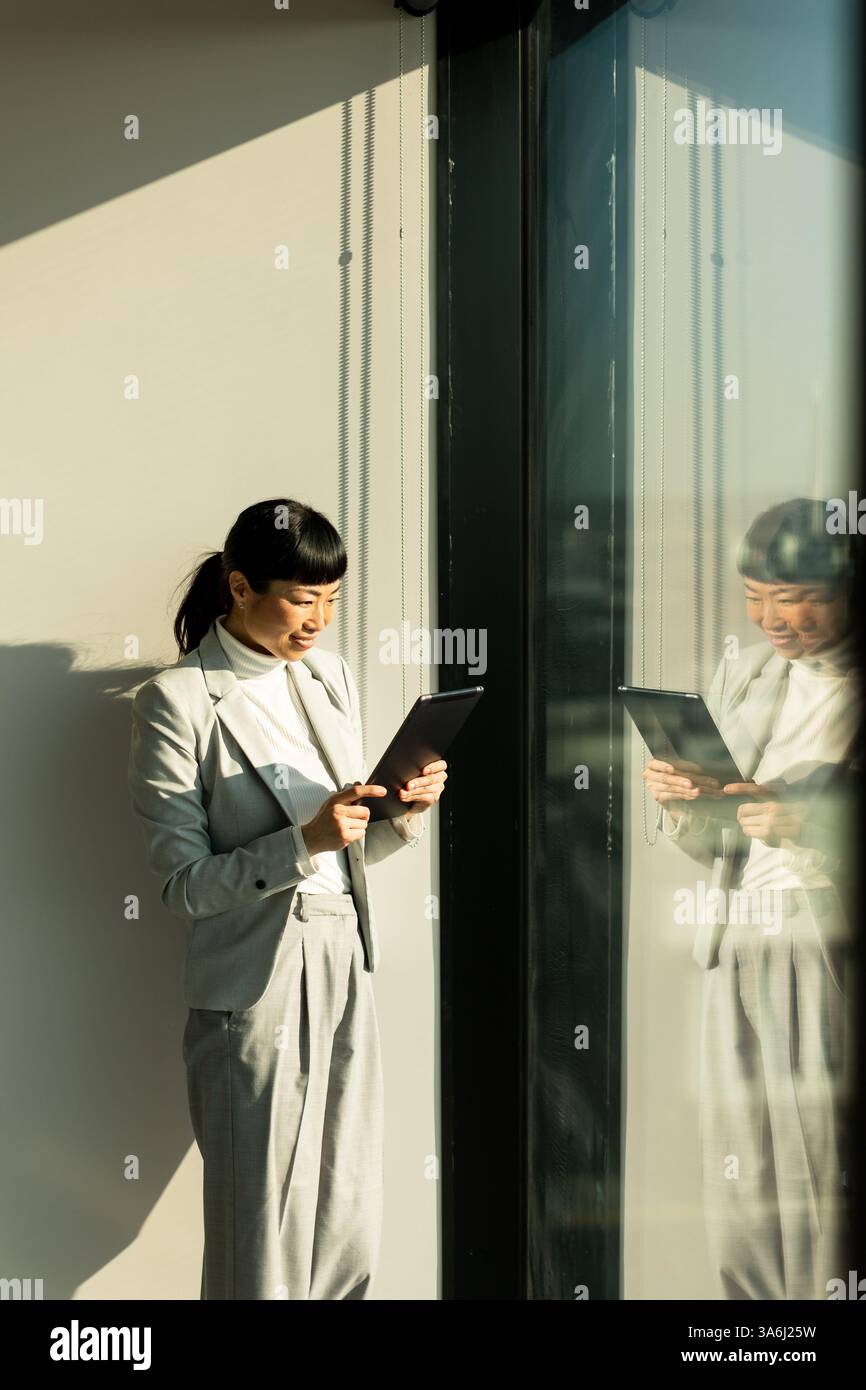 Accanto a una grande finestra in un ufficio moderno, una donna d'affari giapponese usa il suo tablet, assorbito nel suo lavoro. La luce del sole illumina la gioiosa A. Foto Stock