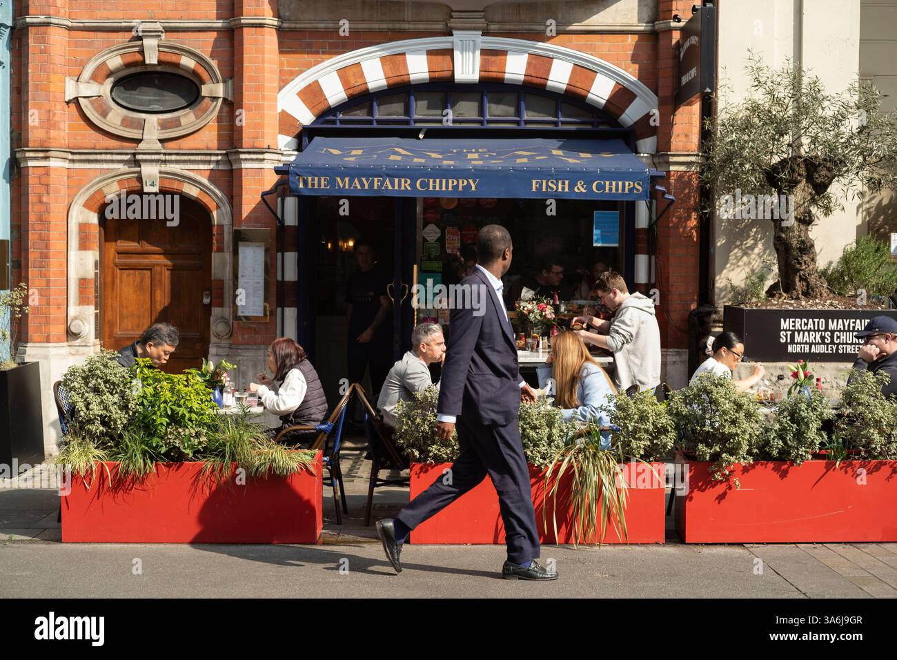 Le persone si siedono fuori dal Mayfair Chippy, in una giornata di primavera soleggiata su North Audley Street, nel cuore di Mayfair, nel centro di Londra, Inghilterra, Regno Unito Foto Stock