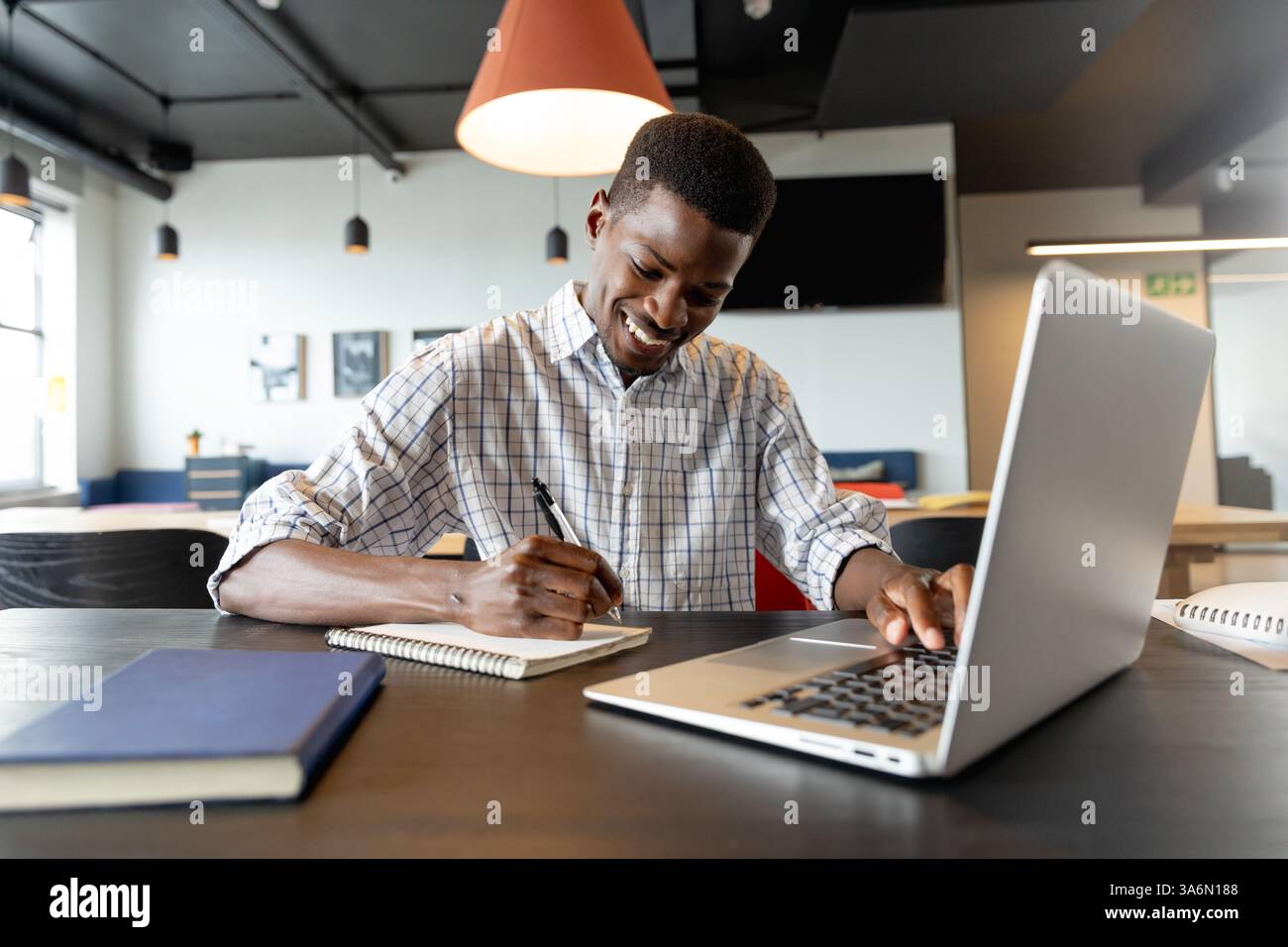 Uomo sorridente che scrive appunti mentre utilizza un computer portatile alla moderna scrivania dell'ufficio Foto Stock