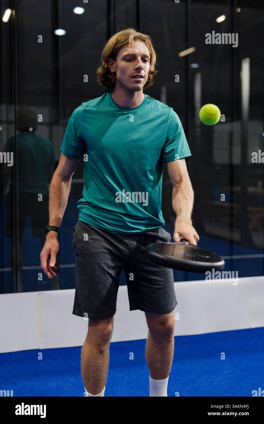 Uomo che pratica il padel tennis in casa, concentrandosi sulla palla con racchetta in mano Foto Stock