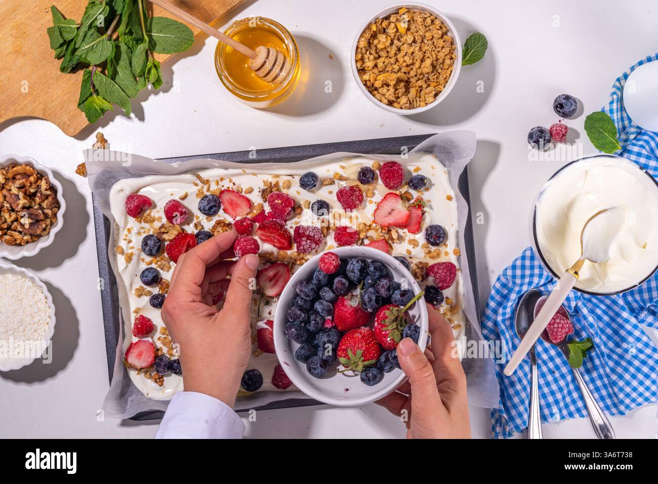 Preparazione della cottura con sfondo di corteccia di yogurt congelato con yogurt greco, bacche fresche, muesli, miele, salsa al cioccolato, con vassoio da forno deco. Vista dall'alto della donna Foto Stock