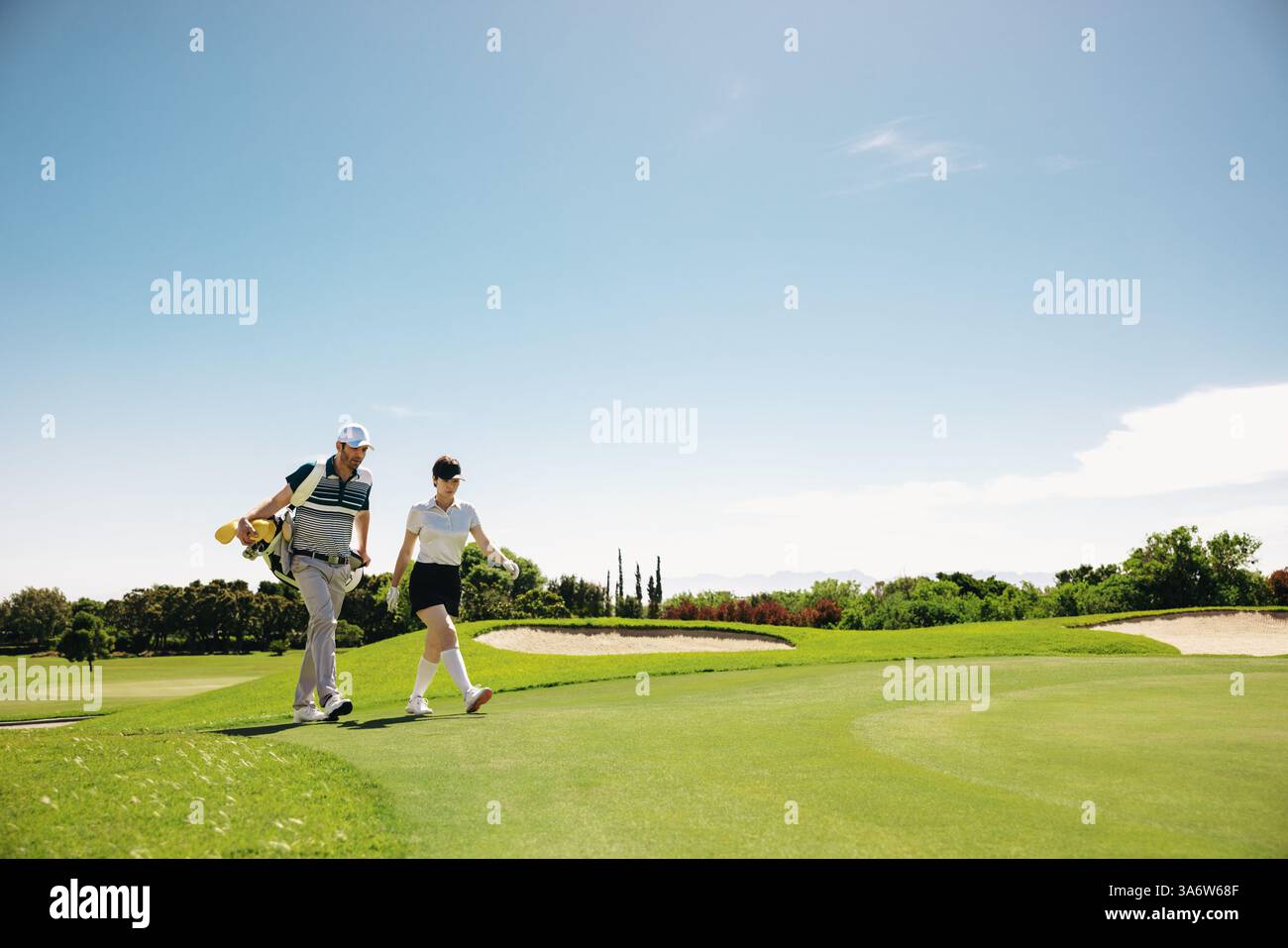 Una golfista e un caddy maschile stanno attraversando il fairway, portando con sé una borsa da golf, su un campo da golf soleggiato durante una partita. Foto Stock