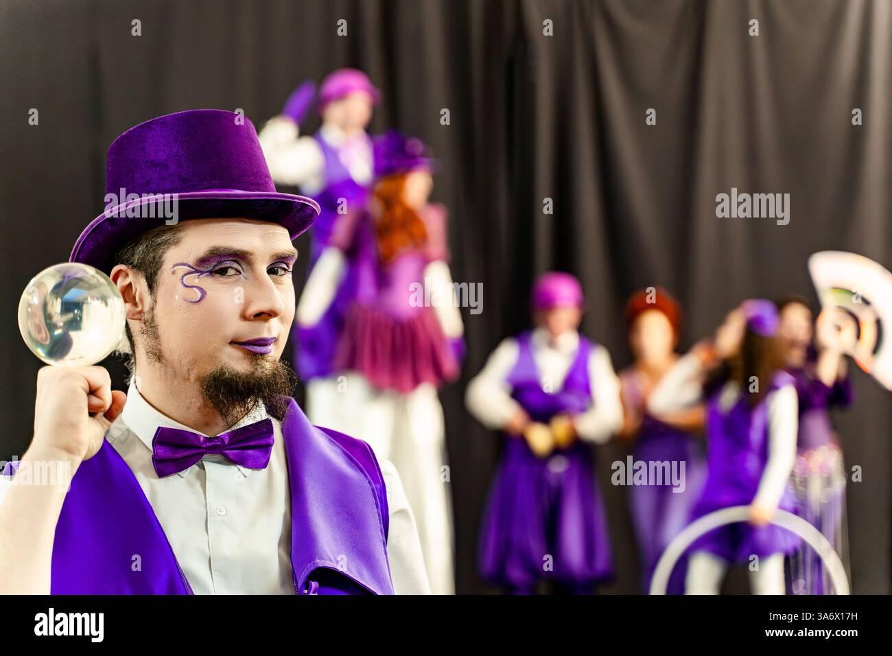 Mago che indossa un costume viola e un cappello con palla di cristallo che si esibisce con una troupe circense sullo sfondo Foto Stock Mago che indossa un costume viola e un cappello con palla di cristallo che si esibisce con una troupe circense sullo sfondo Foto Stock