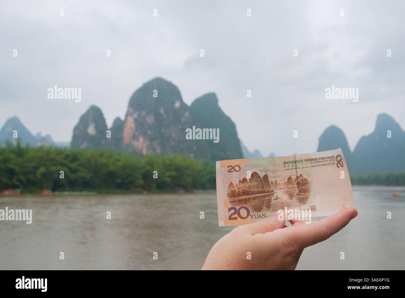 Una mano che tiene una banconota cinese da 20 yuan che corrisponde al paesaggio karstico reale del fiume Li a Guilin in una giornata nuvolosa nel Guangxi, Cina Foto Stock