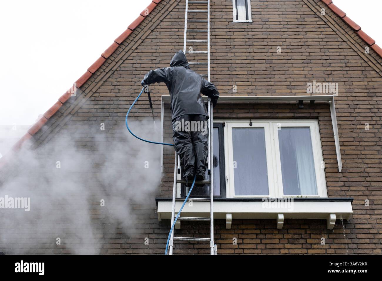 Uomo in tuta impermeabile pulisce la facciata in mattoni sporchi della casa residenziale con un getto d'acqua ad alta pressione. Pulizia ad alta pressione della facciata Foto Stock