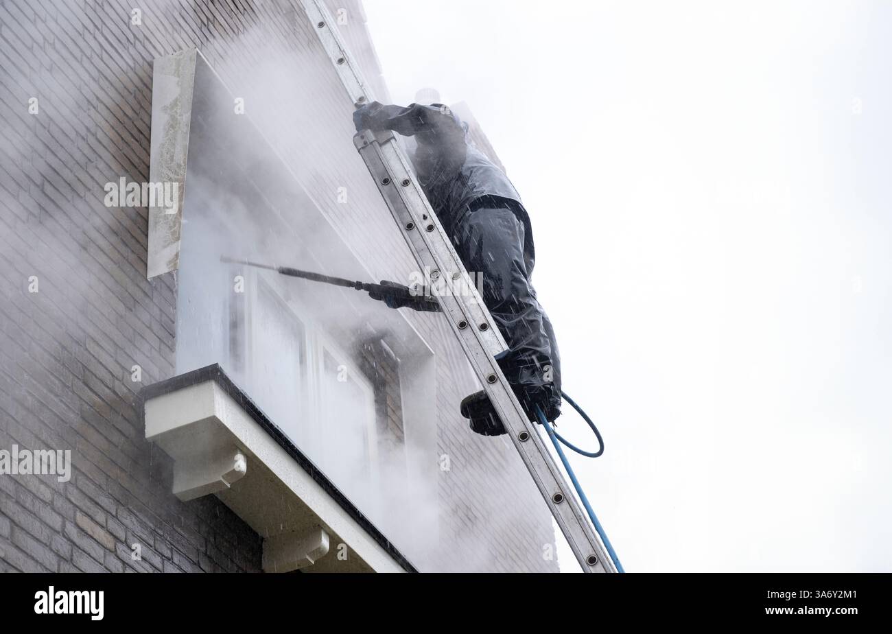 Uomo in tuta impermeabile pulisce la facciata in mattoni sporchi della casa residenziale con un getto d'acqua ad alta pressione. Pulizia ad alta pressione della facciata Foto Stock