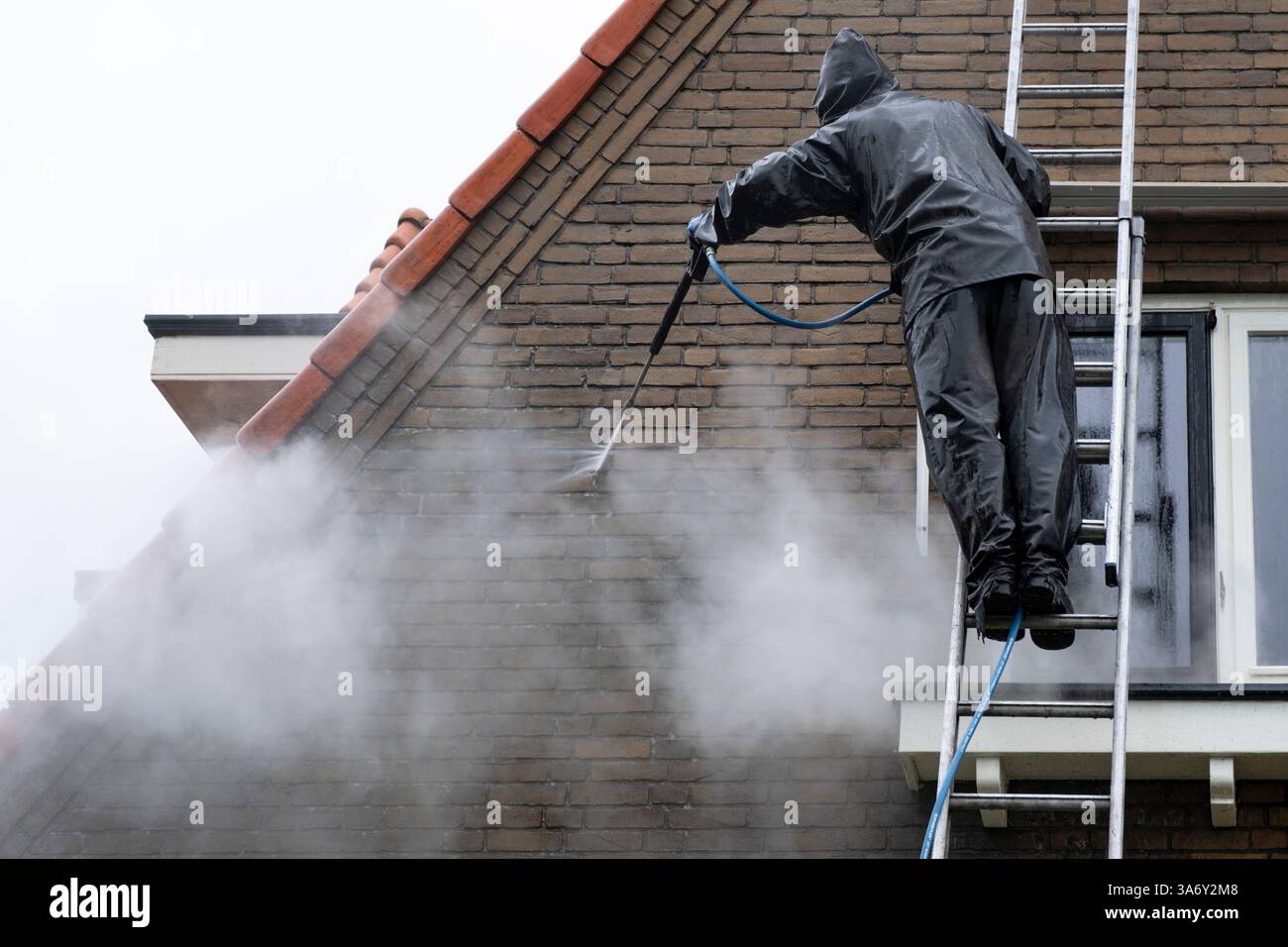 Uomo in tuta impermeabile pulisce la facciata in mattoni sporchi della casa residenziale con un getto d'acqua ad alta pressione. Pulizia ad alta pressione della facciata Foto Stock