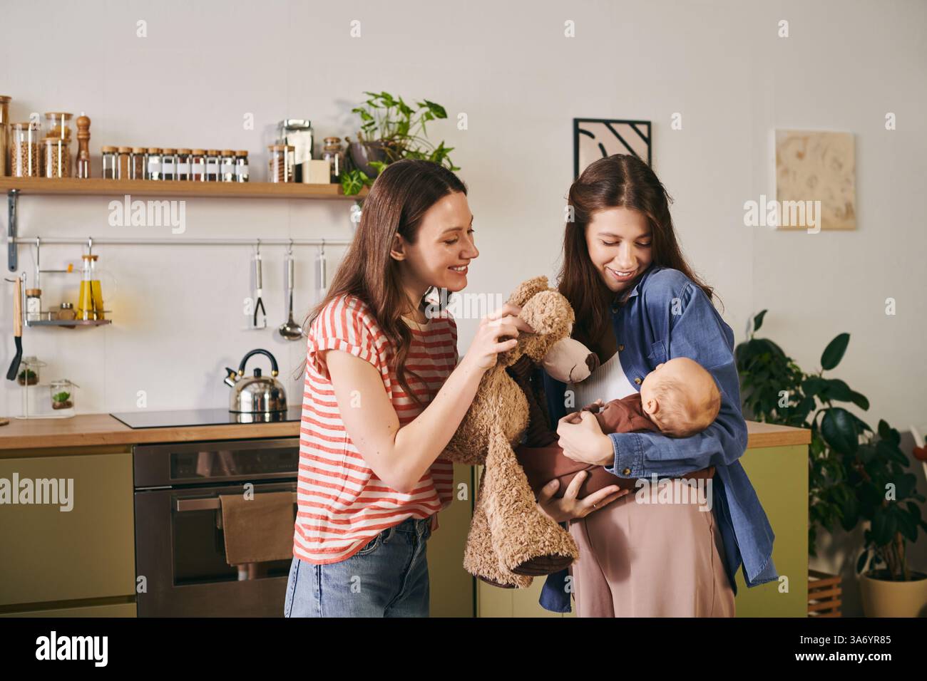 Due madri gioiose si impegnano amorevolmente con il loro bambino in una cucina calda e invitante. Foto Stock Due madri gioiose si impegnano amorevolmente con il loro bambino in una cucina calda e invitante. Foto Stock