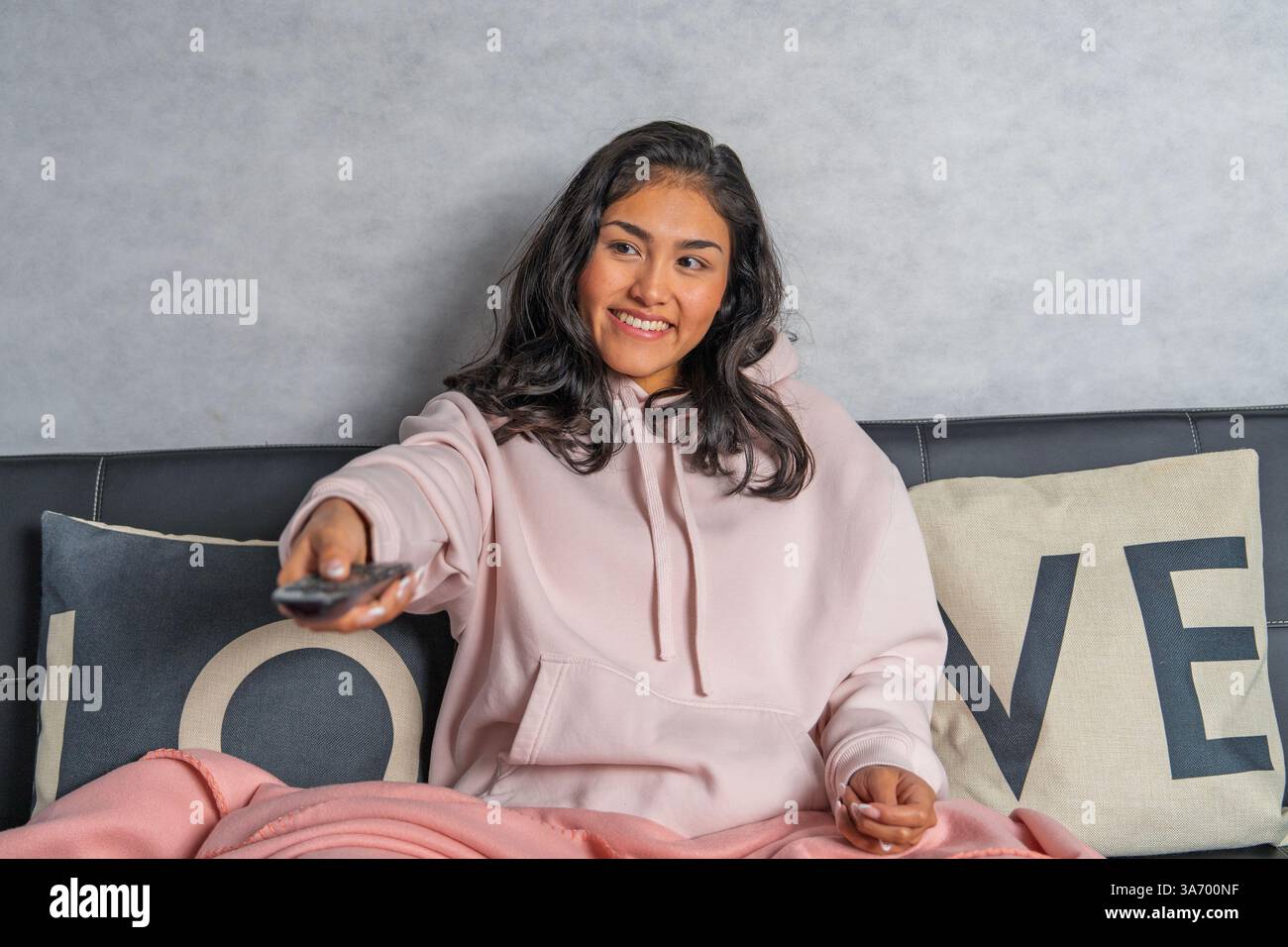 Sorridente giovane donna che guarda il suo programma tv preferito mentre si rilassa su un comodo divano con telecomando in mano Foto Stock