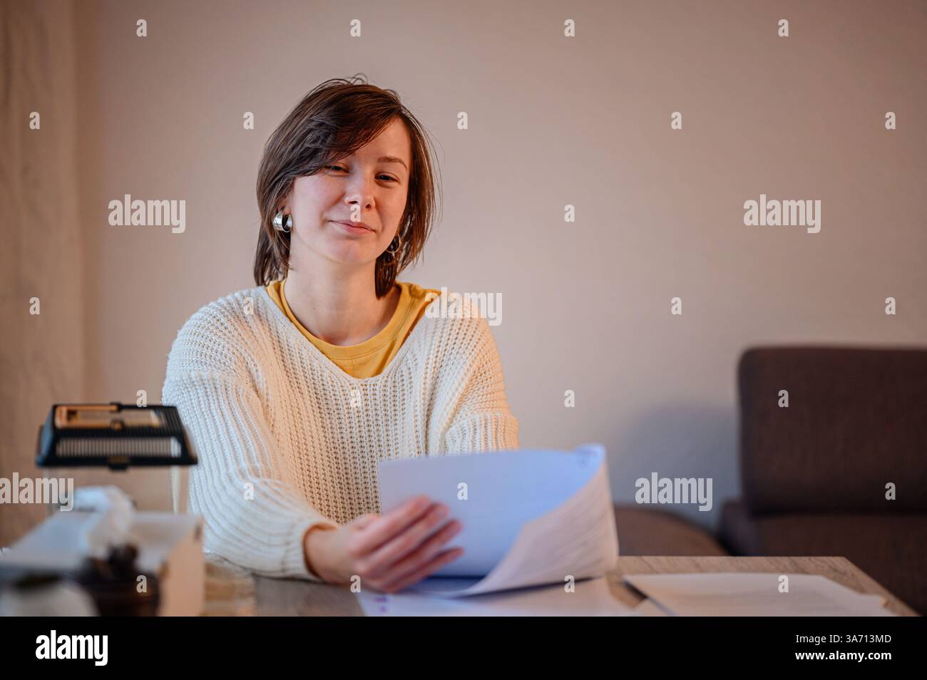 Una donna in un comodo maglione si siede a un tavolo, rivedendo attentamente i documenti. Sembra concentrata, tenendo dei documenti mentre è circondata da materiali di studio An Foto Stock