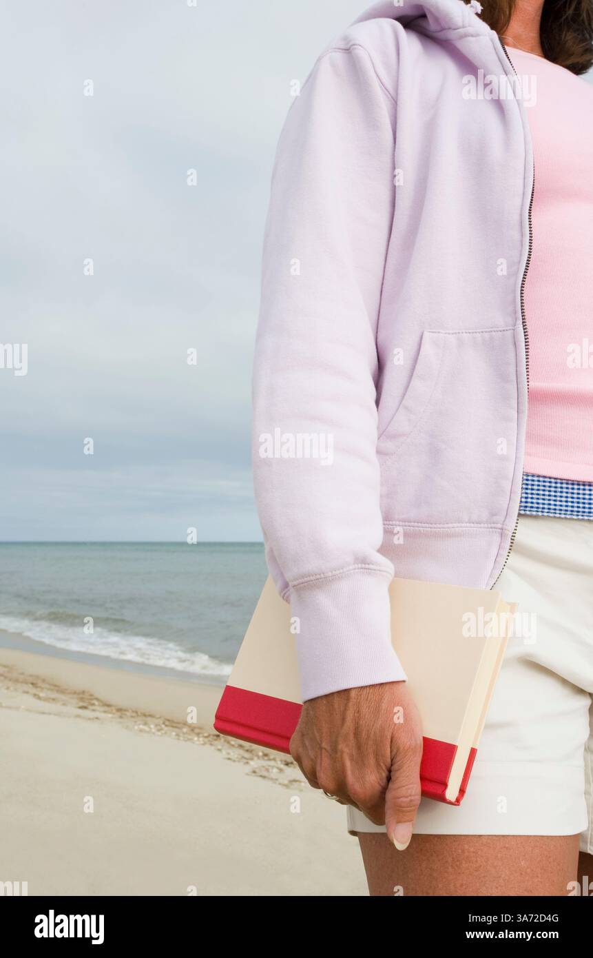 Parte centrale di donna che tiene un libro e infradito bianco sulla spiaggia Foto Stock