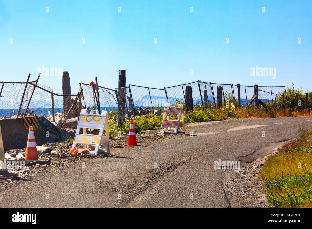 Erosione del litorale, danni a una strada costiera sull'isola di Lummi, macerie e recinzioni rotte sul lato della strada, con coni e cartelli segnaletici. Foto Stock