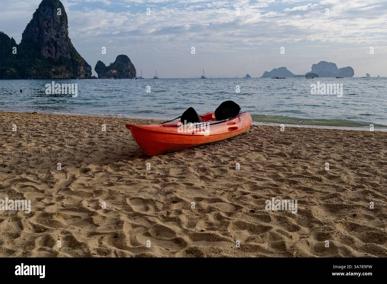 Il kayak rosso poggia sulla sabbia soffice di Tonsai Beach, con scogliere calcaree e barche sparse che punteggiano l'orizzonte Foto Stock
