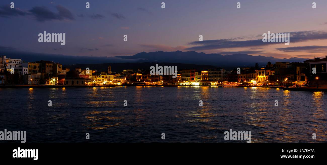 Mentre il crepuscolo si deposita su Chania, Creta, il lungomare si anima con il caldo bagliore di luci che si riflettono sull'acqua. Foto Stock