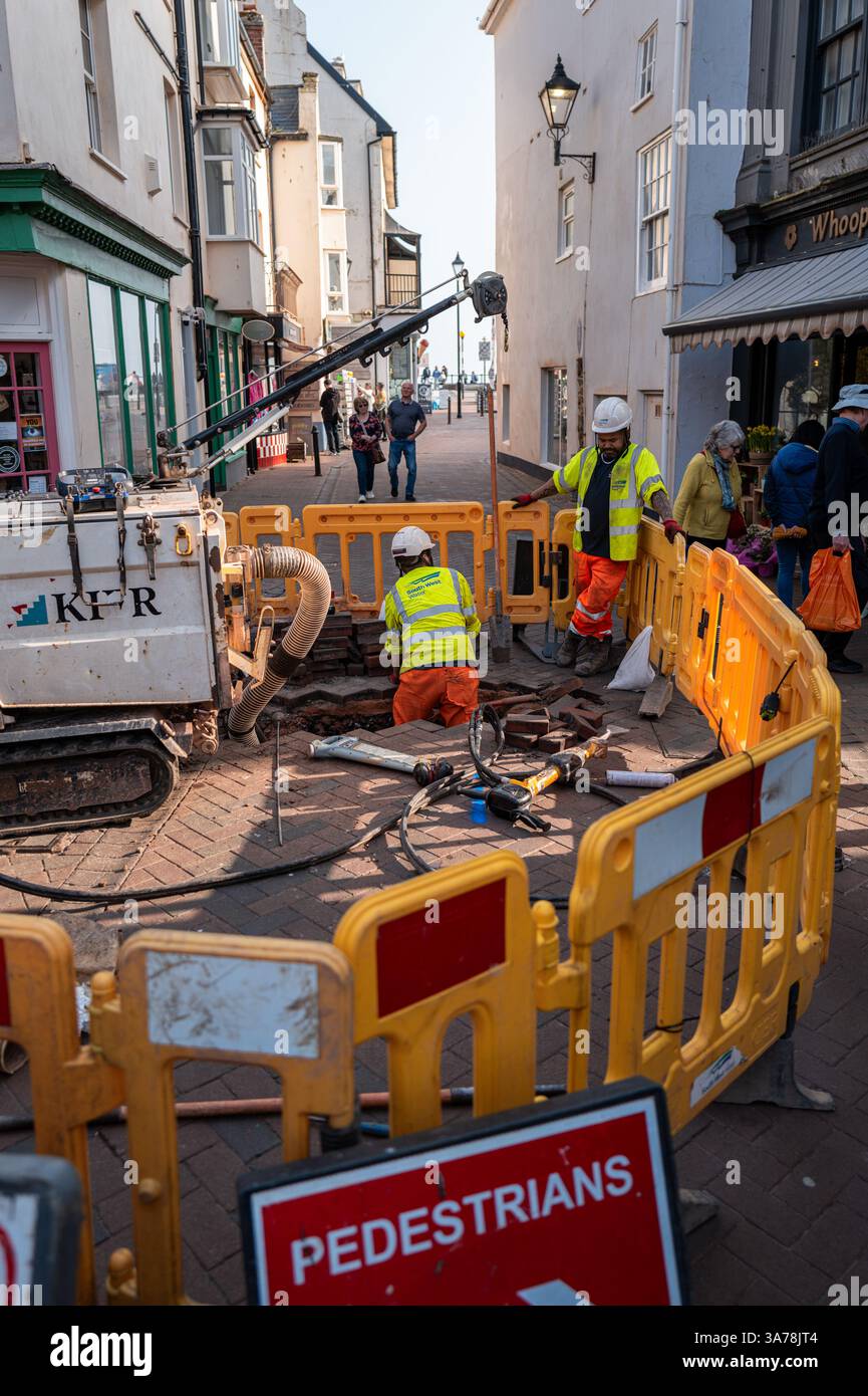 Due addetti alla manutenzione stradale di South West Water circondati da barriere stradali gialle che scavano un buco nella Main Street di Sidmouth, Devon, Regno Unito. Foto Stock