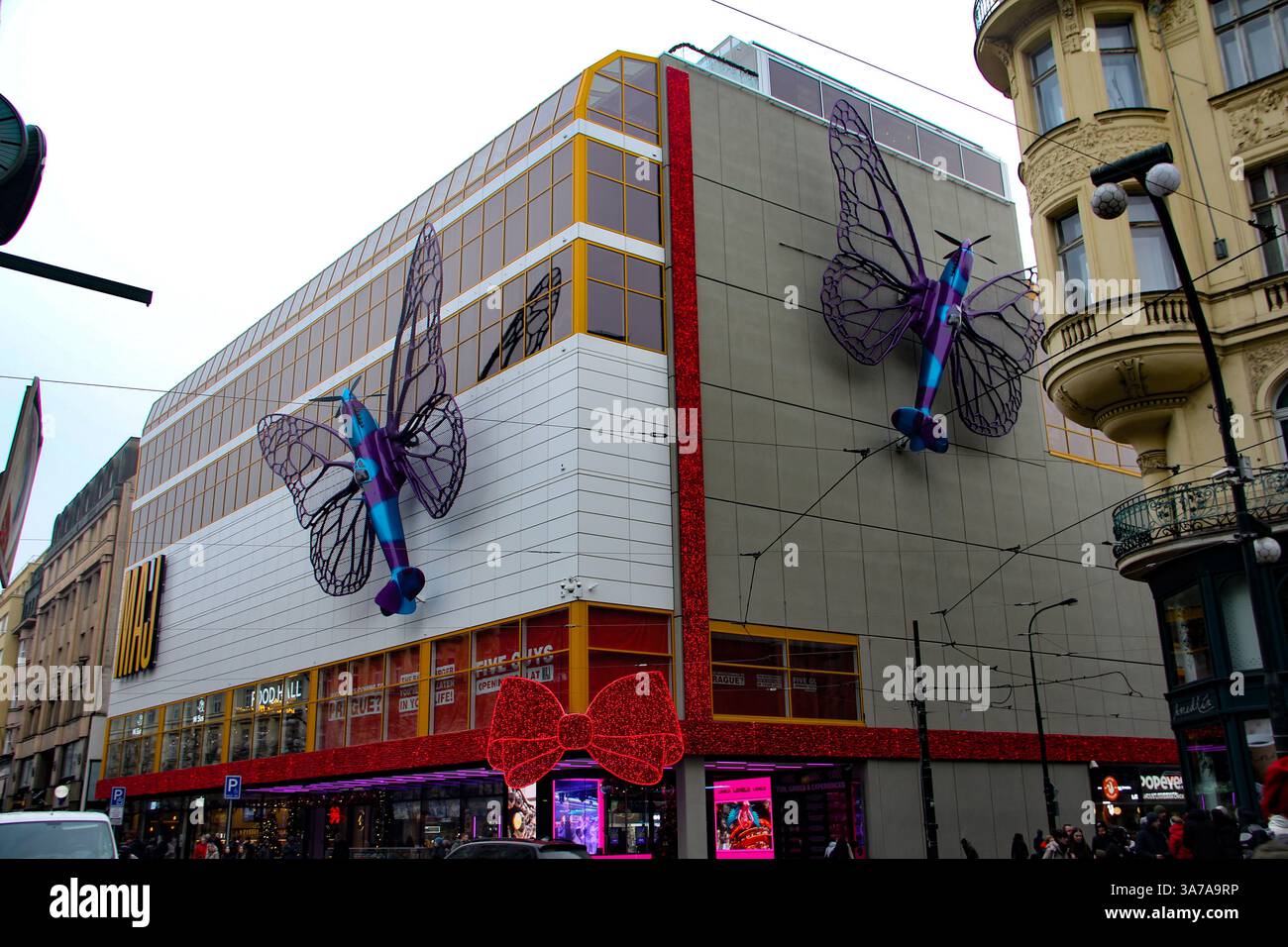 Esterno di un centro commerciale moderno con gigantesche sculture di farfalle viola e una grande decorazione a forma di fiocco rosso in una vivace strada cittadina europea. Foto Stock