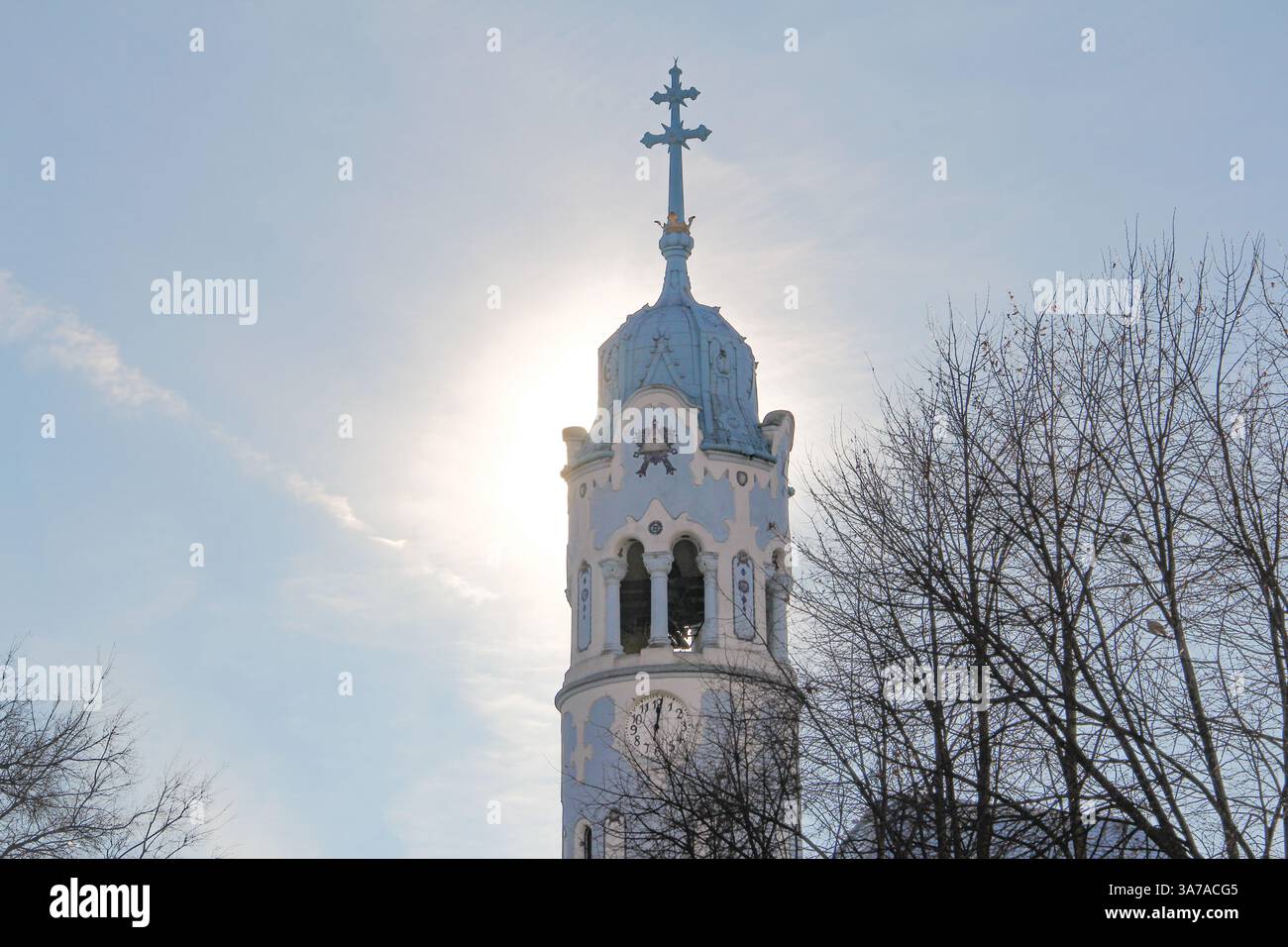 Campanile di chiesa ortodossa blu chiaro con una croce dorata e un orologio, incorniciato da alberi spogli in inverno sotto un cielo blu pallido e soleggiato. Foto Stock
