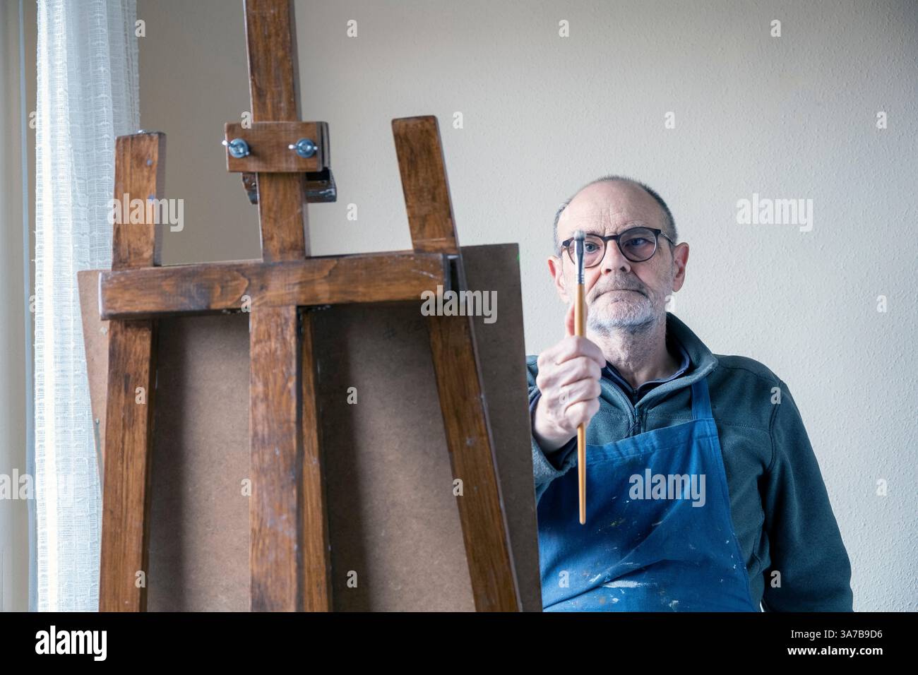 Un vecchio artista maschile, che indossa occhiali e grembiule, dipinge con cura su una tela utilizzando un cavalletto in legno in una stanza ben illuminata. Foto Stock