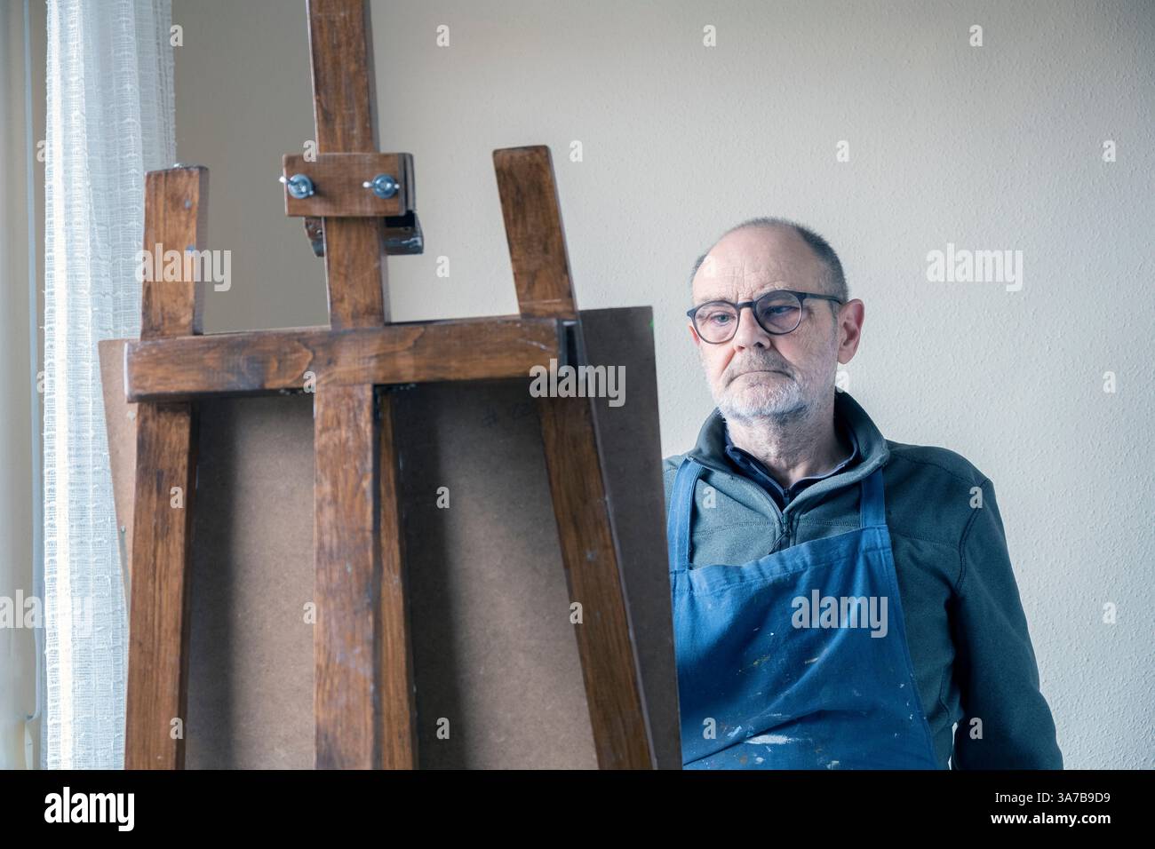 Un vecchio artista maschile, che indossa occhiali e grembiule, dipinge con cura su una tela utilizzando un cavalletto in legno in una stanza ben illuminata. Foto Stock