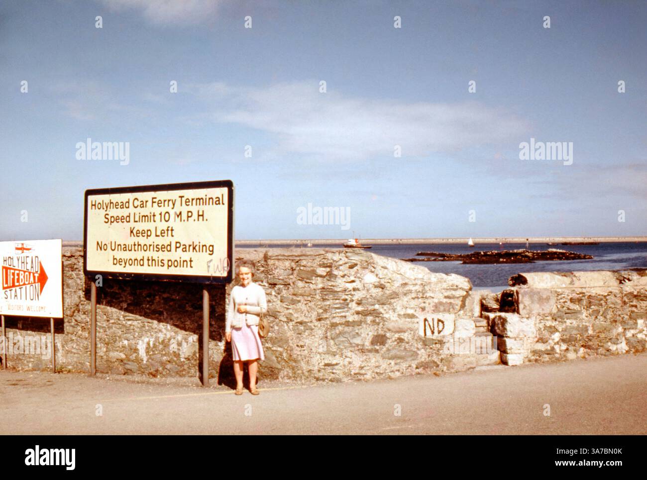 Una donna si trova vicino all'insegna del terminal dei traghetti Holyhead Car Ferry ad Anglesey, nel Galles del Nord, con il Mare d'Irlanda e un traghetto lontano in vista. Ripresa negli anni '1960 su pellicola slide a colori da 35 mm. Foto Stock